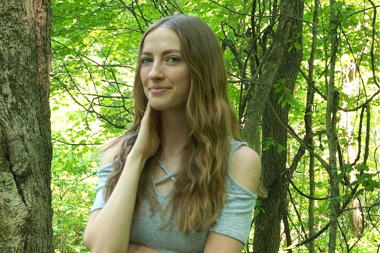 A slender woman with long blonde hair standing in a forest.
